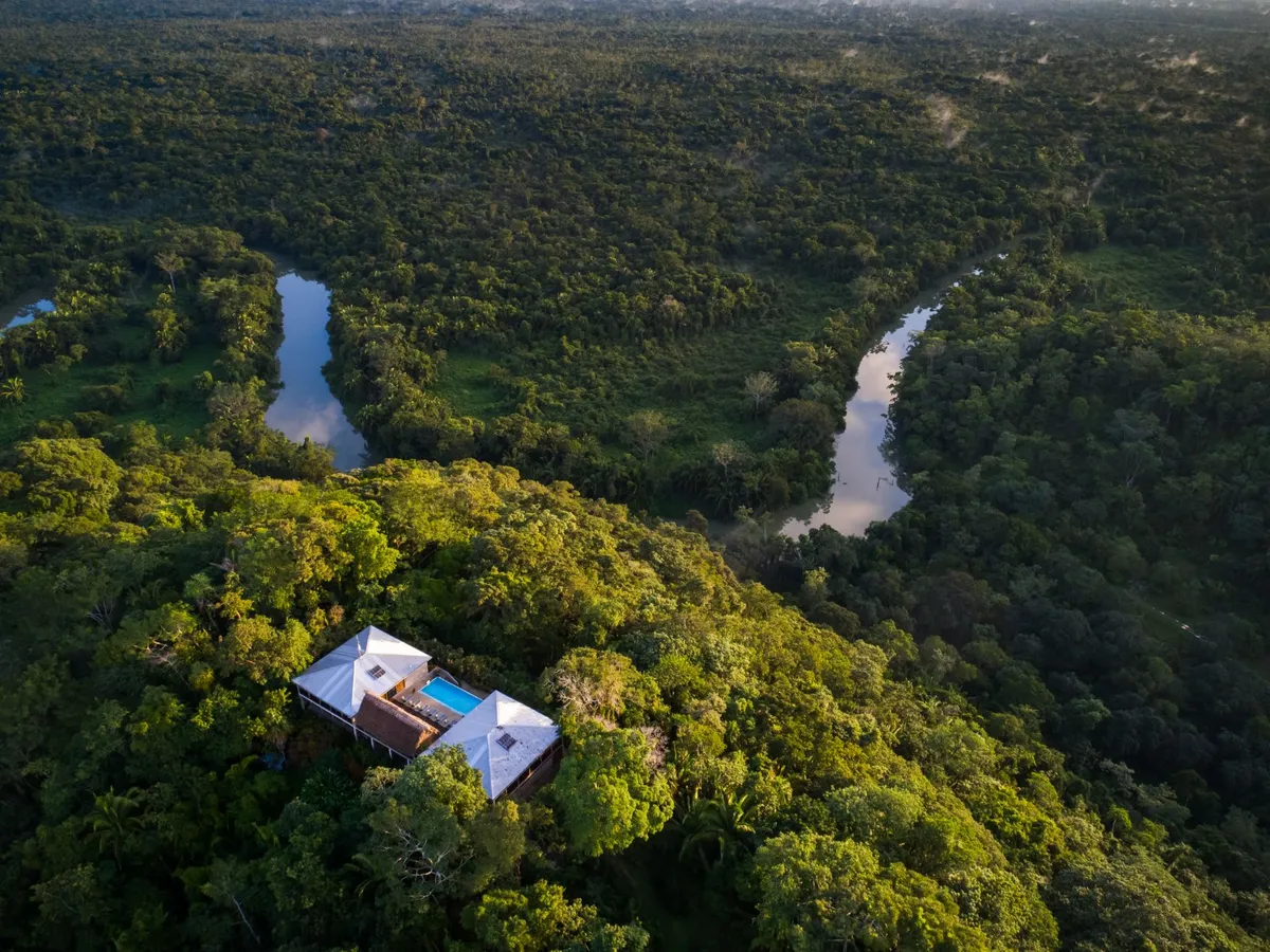 Aerial view of a dense tropical forest with a winding river and a white-roofed house with a blue pool among the trees.