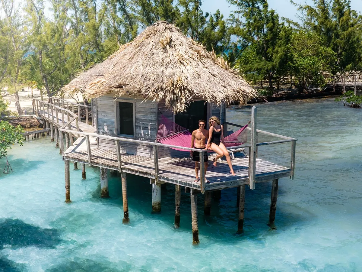 Thatched-roof hut on stilts over turquoise water with a pink hammock and two people relaxing on the deck.