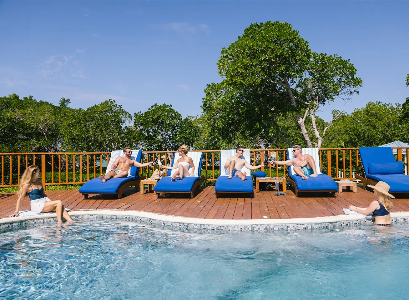 Group of people lounging on blue pool chairs around a deck, chatting and toasting by a clear pool on a sunny day.