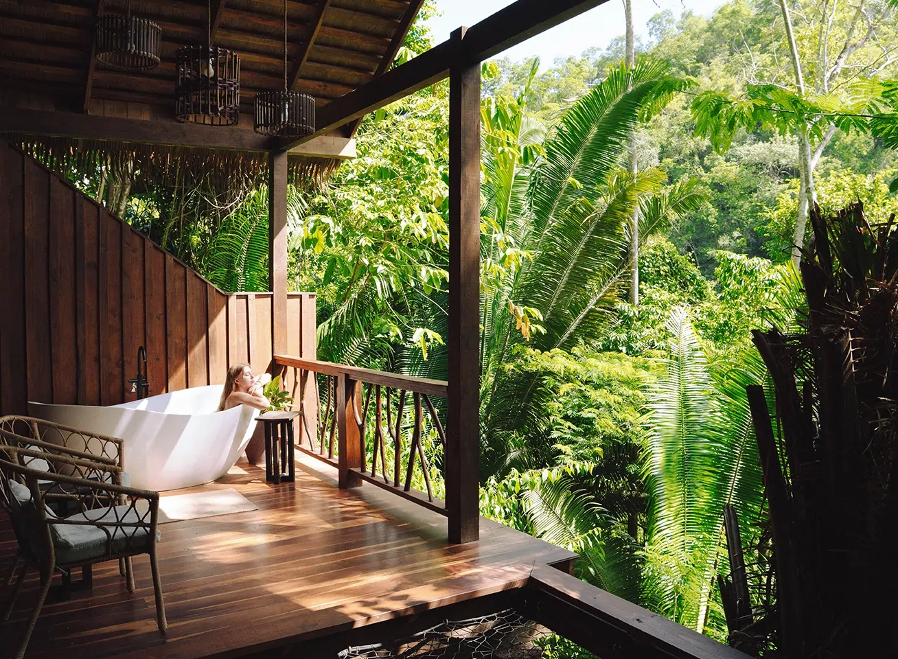 Woman relaxing in a white freestanding tub on a wooden jungle deck, surrounded by dense tropical foliage.
