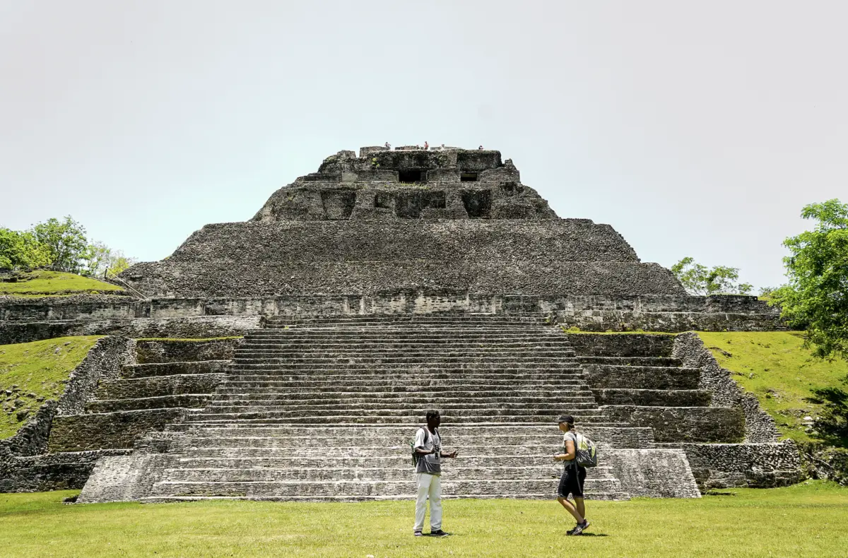 Ancient stepped stone pyramid with two people standing on the grass in front.