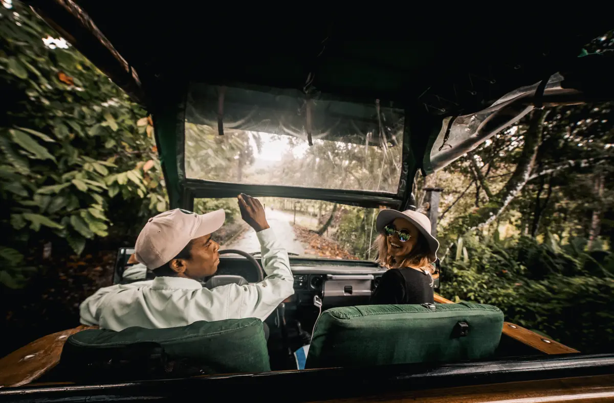 Two friends ride in an open-top jeep along a leafy forest road; one driver waves while the passenger smiles at the camera with a hat and sunglasses.
