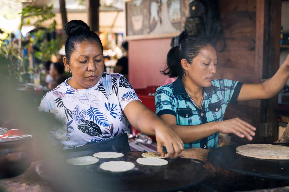 Two women cook small tortillas on hot griddles at a market or outdoor kitchen.