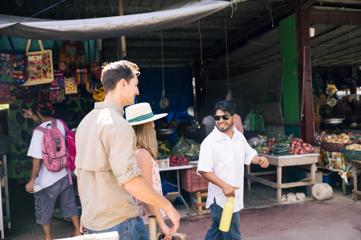 Tourist couple chats with a market vendor at an open-air produce stall, a yellow bottle in the vendor's hand.