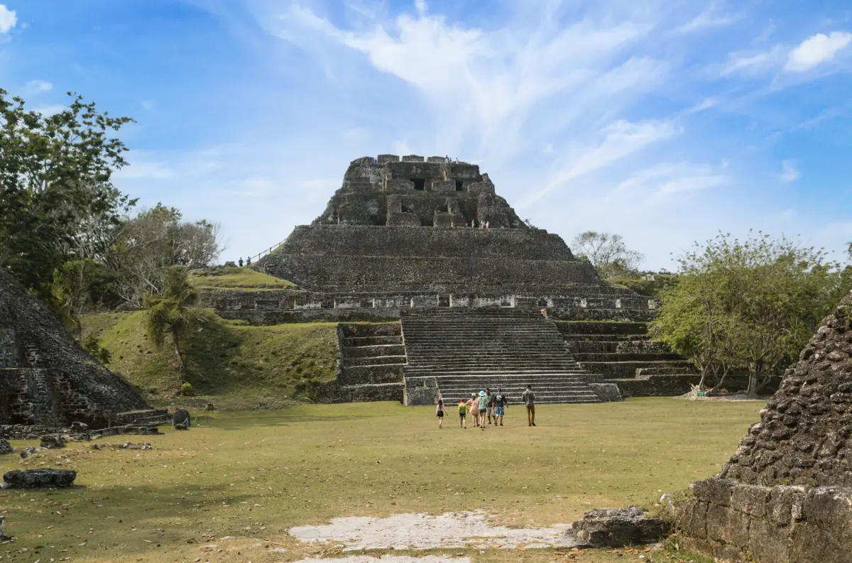 Ancient stepped stone temple with a long stairway in a grassy plaza; a small group of tourists approaches the steps.
